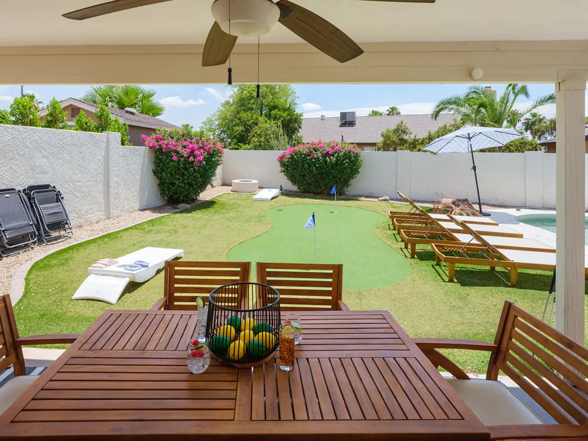 An exterior shot showing a well-manicured backyard featuring a dining table with a fruit basket, a putting green, sun loungers, umbrella, and colorful flowers. The scene evokes the idea of outdoor play, golf practice, and leisurely gatherings.