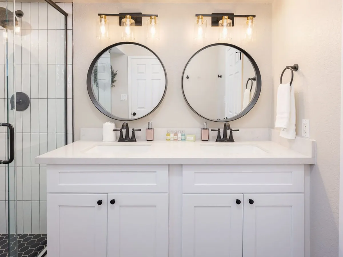 A modern bathroom with dual circular mirrors, sleek light fixtures, a white vanity, and a glass shower enclosure. Morning sunlight reflects off surfaces, creating a refreshing and spa-like atmosphere.