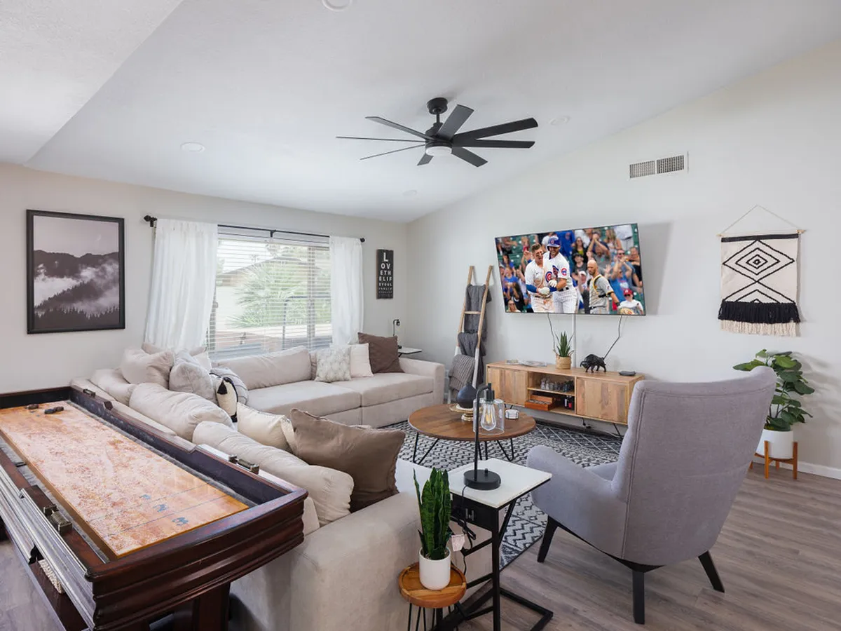 A vibrant, open-concept living space featuring a large sectional sofa facing an oversized television. The room seamlessly flows into a game area with a pool table, surrounded by wall art and stylish decor. The area is brightly lit and clearly designed for relaxation and socializing.