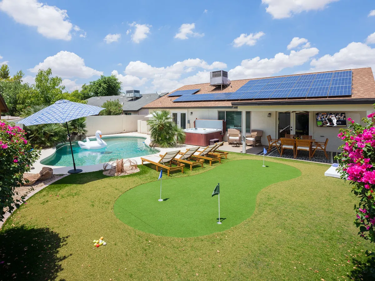 A vibrant backyard scene featuring a sparkling swimming pool, putting green, inviting hot tub, several lounge chairs, a covered patio with dining furniture, and decorative flowers. The space highlights multiple activity zones, from relaxing in the sun to enjoying mini-golf or an alfresco meal under cover.