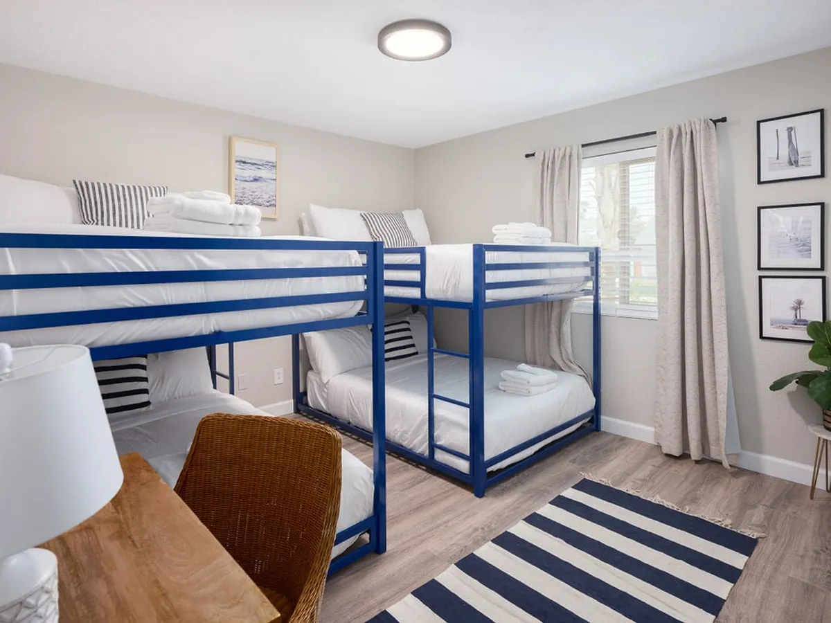 A cheerful bunk room featuring two sets of bunk beds, a desk area for reading or studying, playful wall art, and a window with curtains that lets in natural light. The ambiance is communal and inviting—well-suited for children or group travelers.