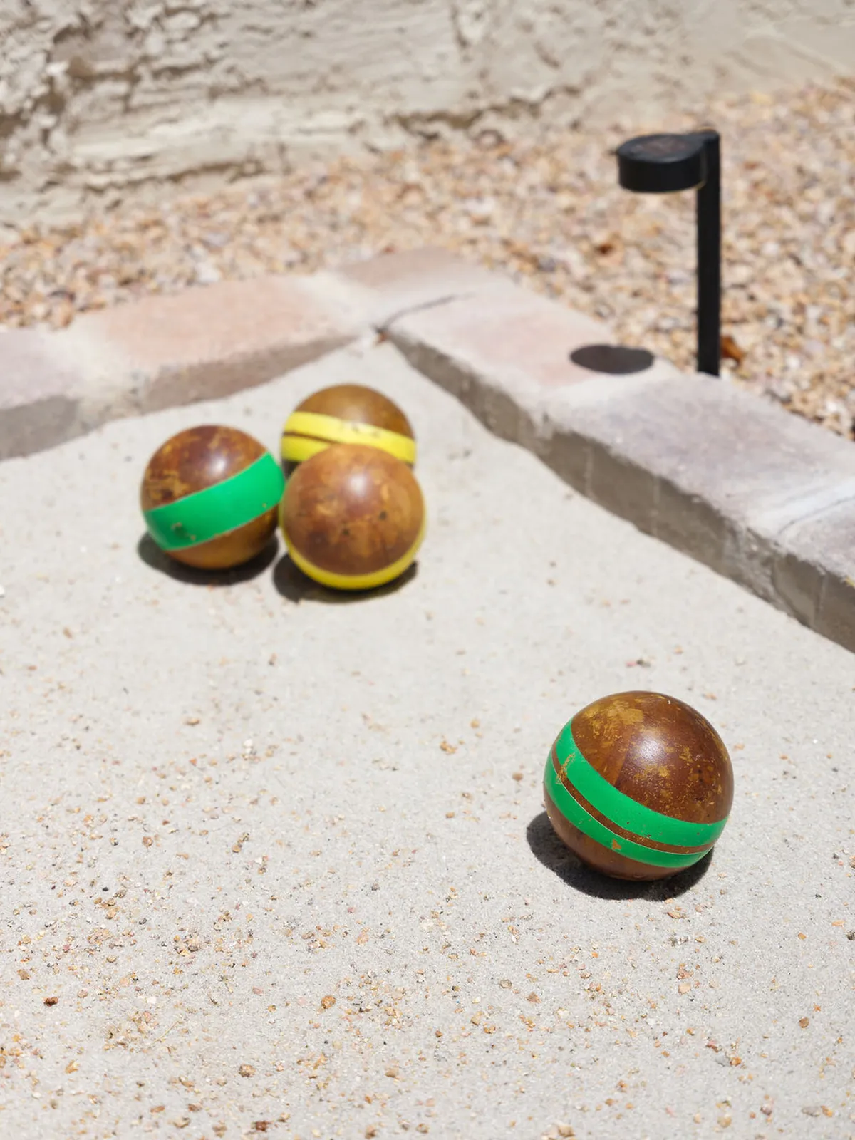 A close-up of a bocce ball court with colorful balls arranged on the sandy surface, bordered by stones. The sunny outdoor environment suggests a setting for friendly competition and socializing.