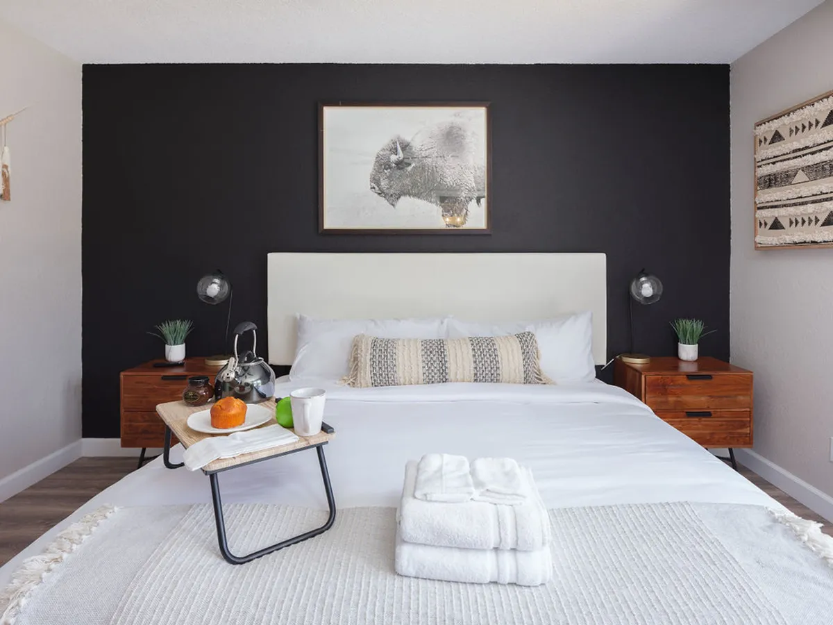 A king bedroom with a soothing black accent wall, crisp white linens, wooden nightstands, and a striking buffalo artwork above the bed. The space is bathed in natural light, with a breakfast tray on the bed suggesting a serene morning routine.