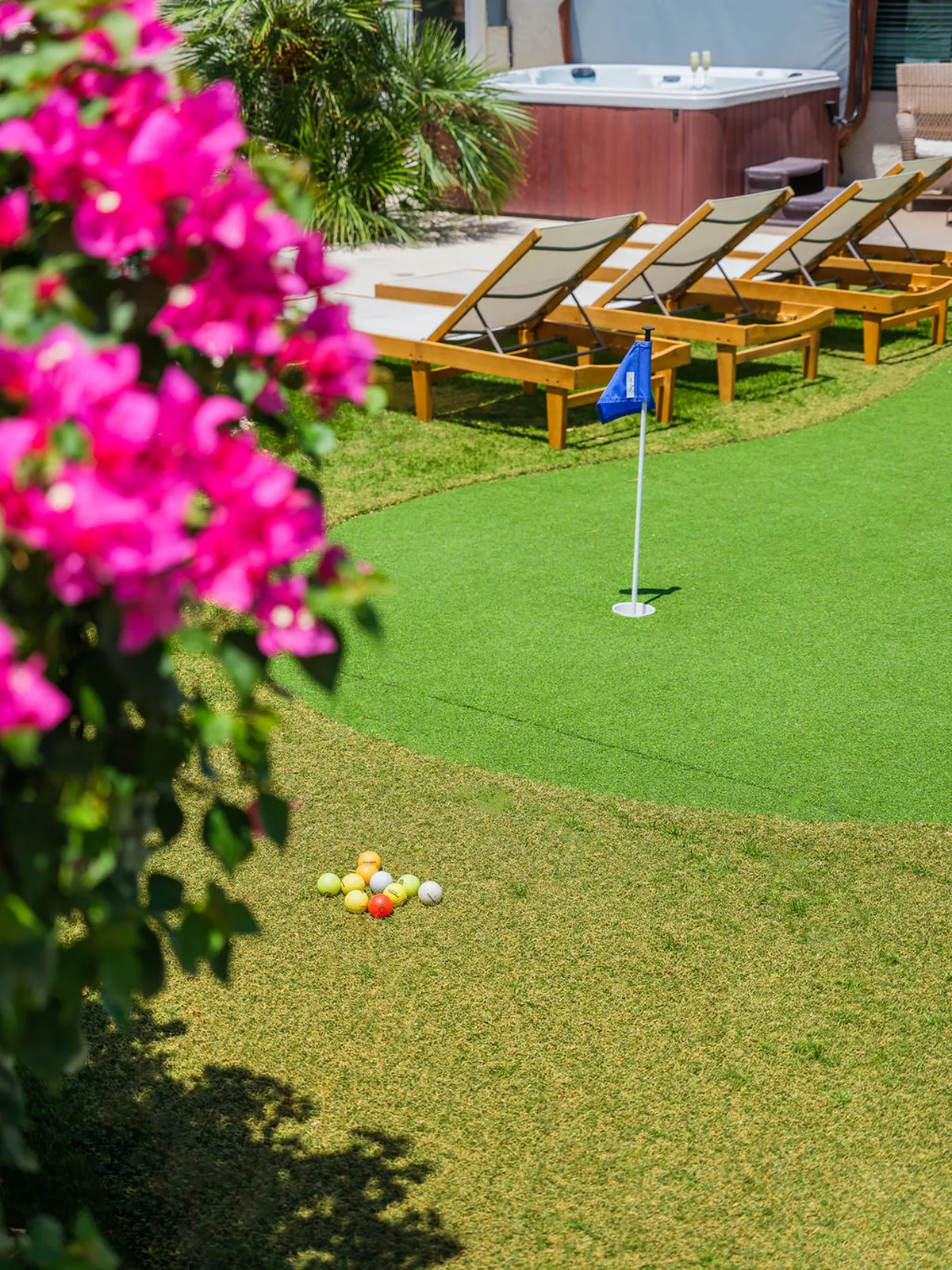 A view of the private backyard putting green, complete with colorful golf balls, adjacent to a pool area with lounge chairs and vibrant flowers. The scene captures guests practicing their short game and relaxing in a lush, resort-style yard.
